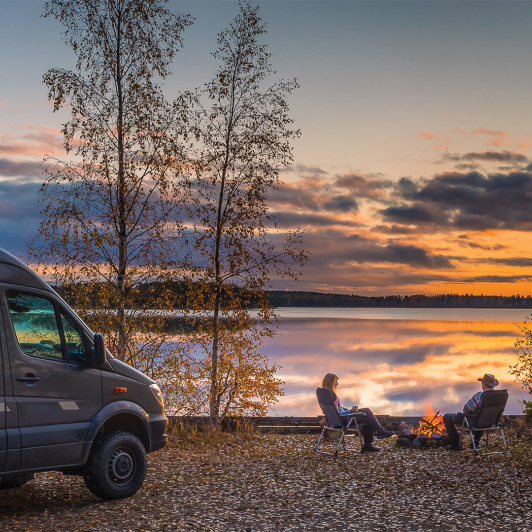 Bild på ett par som sitter vid en husbil i varsin campingstol framför en brasa, vid en sjö i solnedgång.