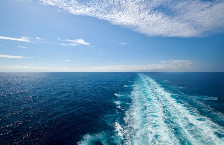 View of the open ocean with a clear blue sky and a white foamy wake trail left by a boat or ship moving through the water.