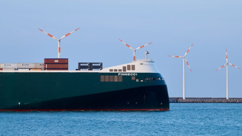 Large cargo ship named Finneco 1 sailing on calm blue water with several wind turbines in the background against a clear sky.