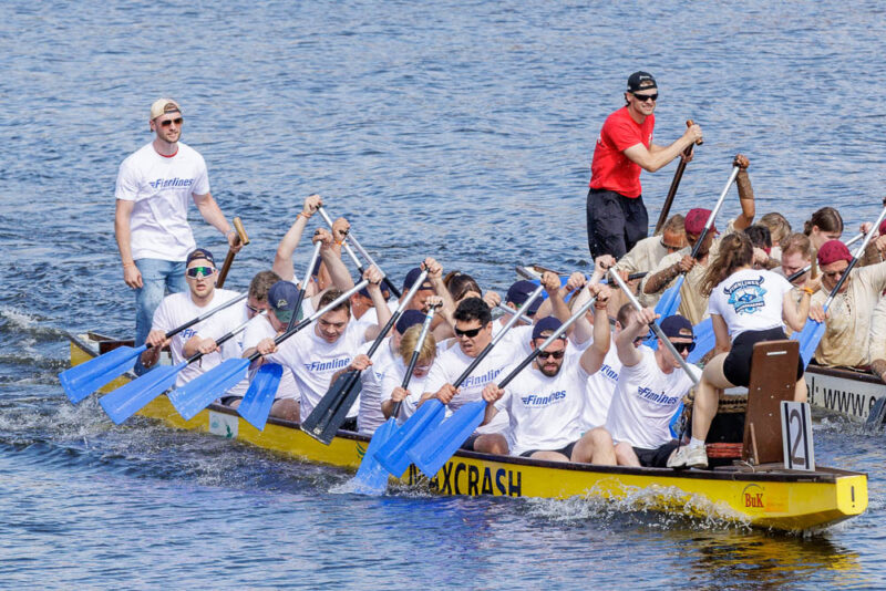 A team of paddlers in a yellow dragon boat wearing white shirts and sunglasses, rowing vigorously on calm blue water during the Finnlines Icebreaker dragon boat race.