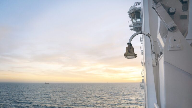 View from the side of a white ship at sunrise with a calm, pastel-colored sea and sky, a deck lamp and ship superstructure in the foreground on the right, and a small vessel on the distant horizon.