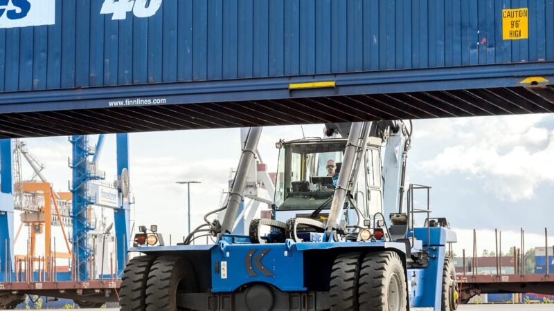 Front view of a large blue container handler (reach stacker) with heavy tires and a lifting spreader holding a shipping container at a busy port, with gantry cranes and stacked containers in the background under a cloudy sky.