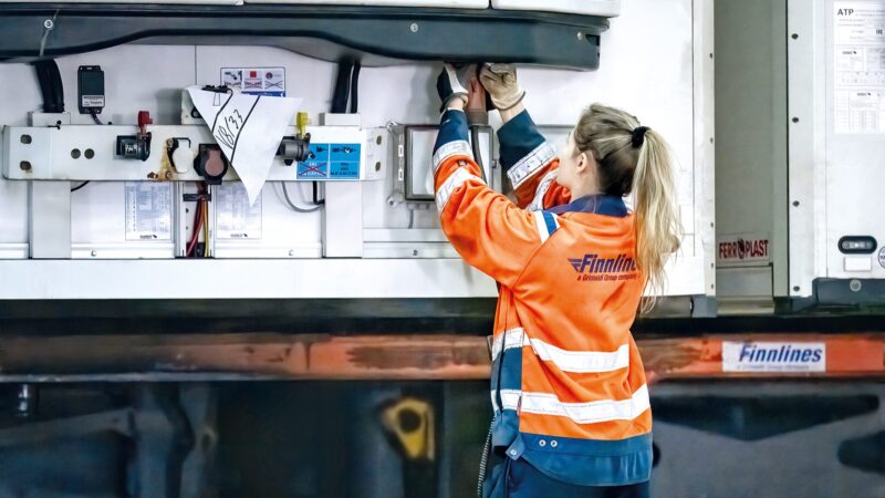 Person with hair tied back wearing an orange high-visibility Finnlines jacket and blue trousers reaches up into the open rear of a trailer to adjust equipment or secure cargo, with control panels and paperwork visible on the trailer wall.
