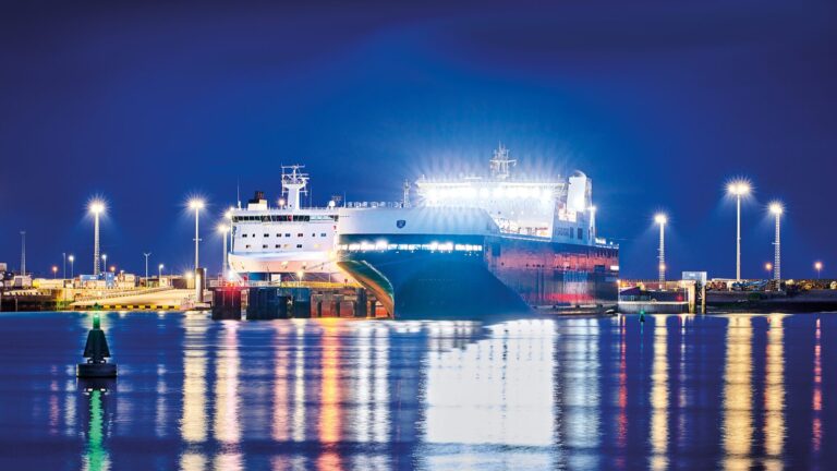 Night view of the FinnEco I ferry docked at Zeebrugge, brightly lit by floodlights with pier lights and long shimmering reflections on calm water and a small buoy in the foreground