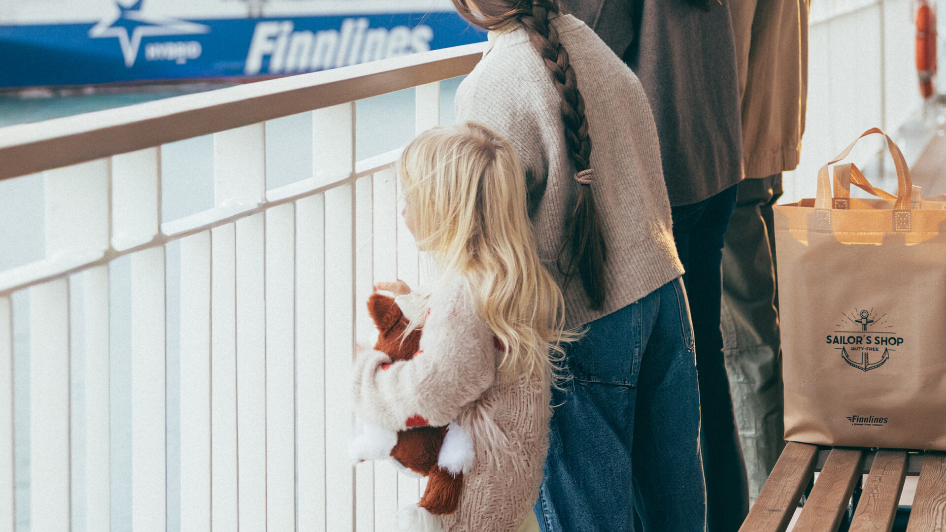 Bild på en familj och liten flicka i fokus som håller i ett gosedjur. På bänken bakom står en påse från Sailor's shop.